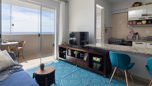 Una sala de estar moderna con televisor, sofá y sillones, un balcón con vistas, y una zona de cocina.
