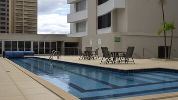 Piscina com borda azul, rodeada por um deck revestido de azulejos, com algumas cadeiras e uma espreguiçadeira azul.