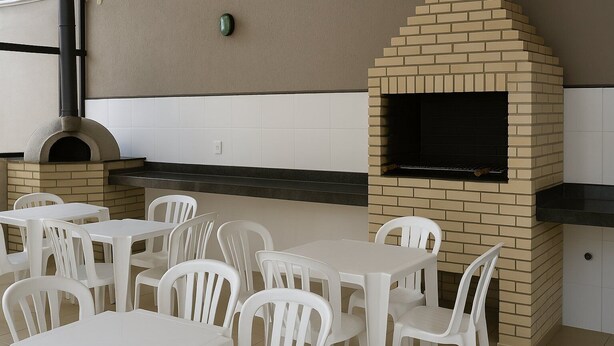 Un patio al aire libre con horno de ladrillo y parrilla, sillas y mesas de plástico blanco.