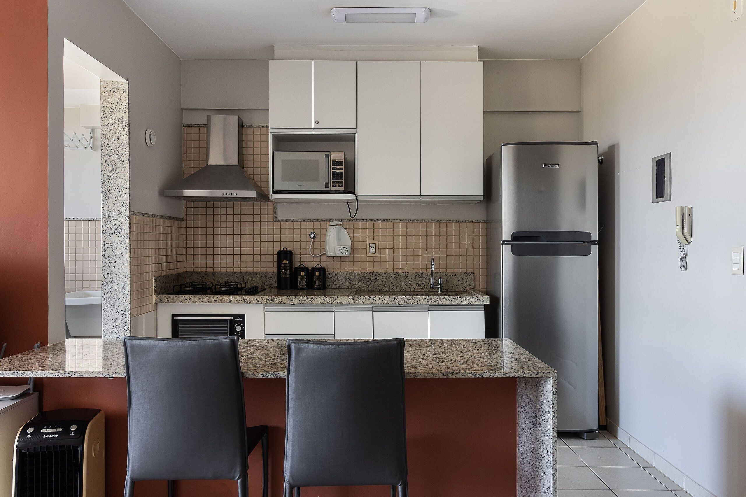 Una cocina moderna con armarios blancos, un refrigerador de acero inoxidable, un microondas y una estufa a gas. Hay dos taburetes de bar en la encimera.