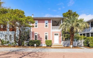 A two-story pink building with a balcony, surrounded by trees and a palm tree.