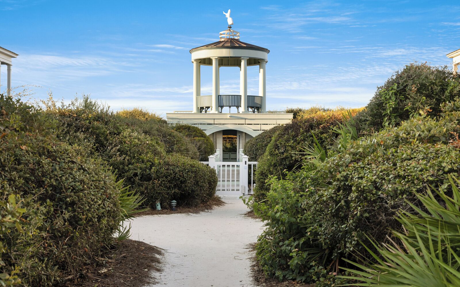 A gazebo with a white exterior and a central seating area, surrounded by dense greenery and a pathway leading to it.