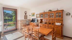 A dining area with a wooden table and chairs, a kitchen with wooden cabinets, a television, and a view of greenery through a sliding door.
