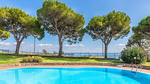 A swimming pool with a clear view of the sea, surrounded by trees and a stone wall.