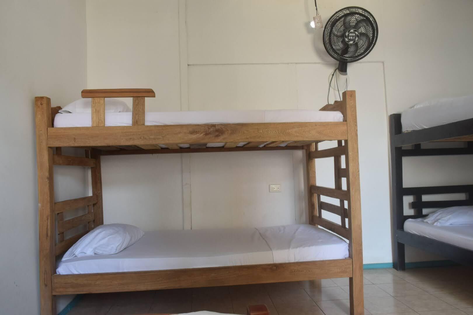 A bunk bed with a wooden frame, white bedding, and a small table on the top bunk. A fan is mounted on the wall above the bed.