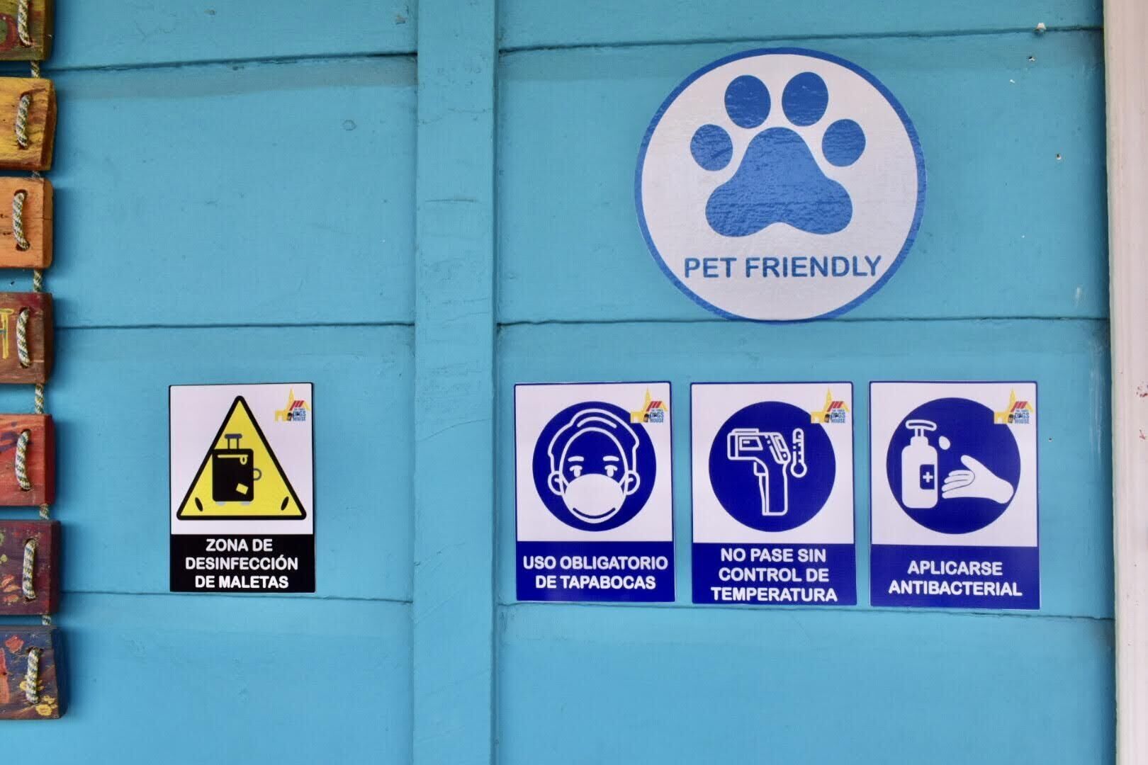 Pared azul con señales que indican: admite mascotas, zona de desinfección, uso obligatorio de mascarilla, control de temperatura y dispensador de gel antibacterial.
