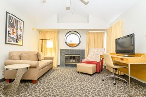 Habitación de hotel con sofá, sillón, escritorio, televisor, chimenea y cuadros en la pared.