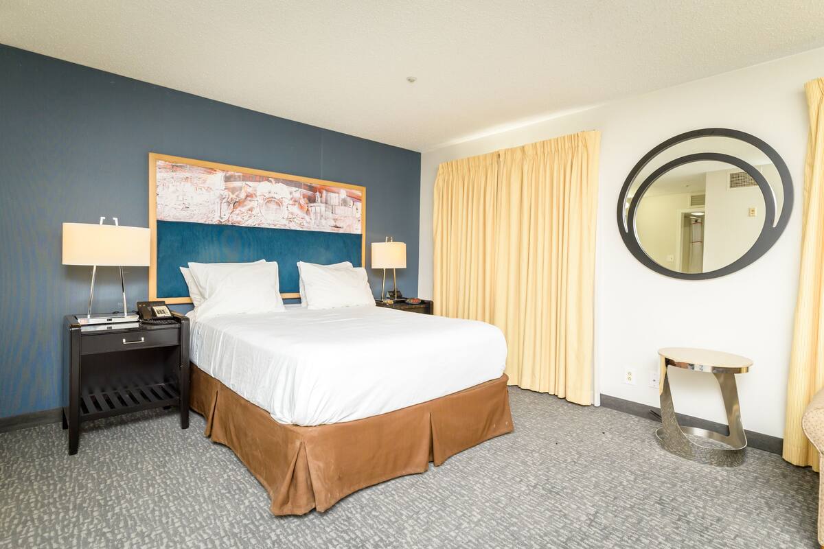 A hotel room with a large bed, two bedside tables with lamps, a round mirror, and a view of the hallway.