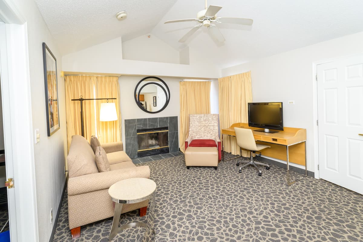 A hotel room with a sofa, armchair, desk, television, and a fireplace.