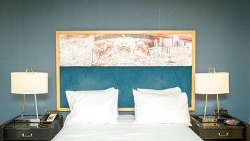 A hotel room with a bed, two bedside tables with lamps, a telephone, and a large cityscape painting on the wall.