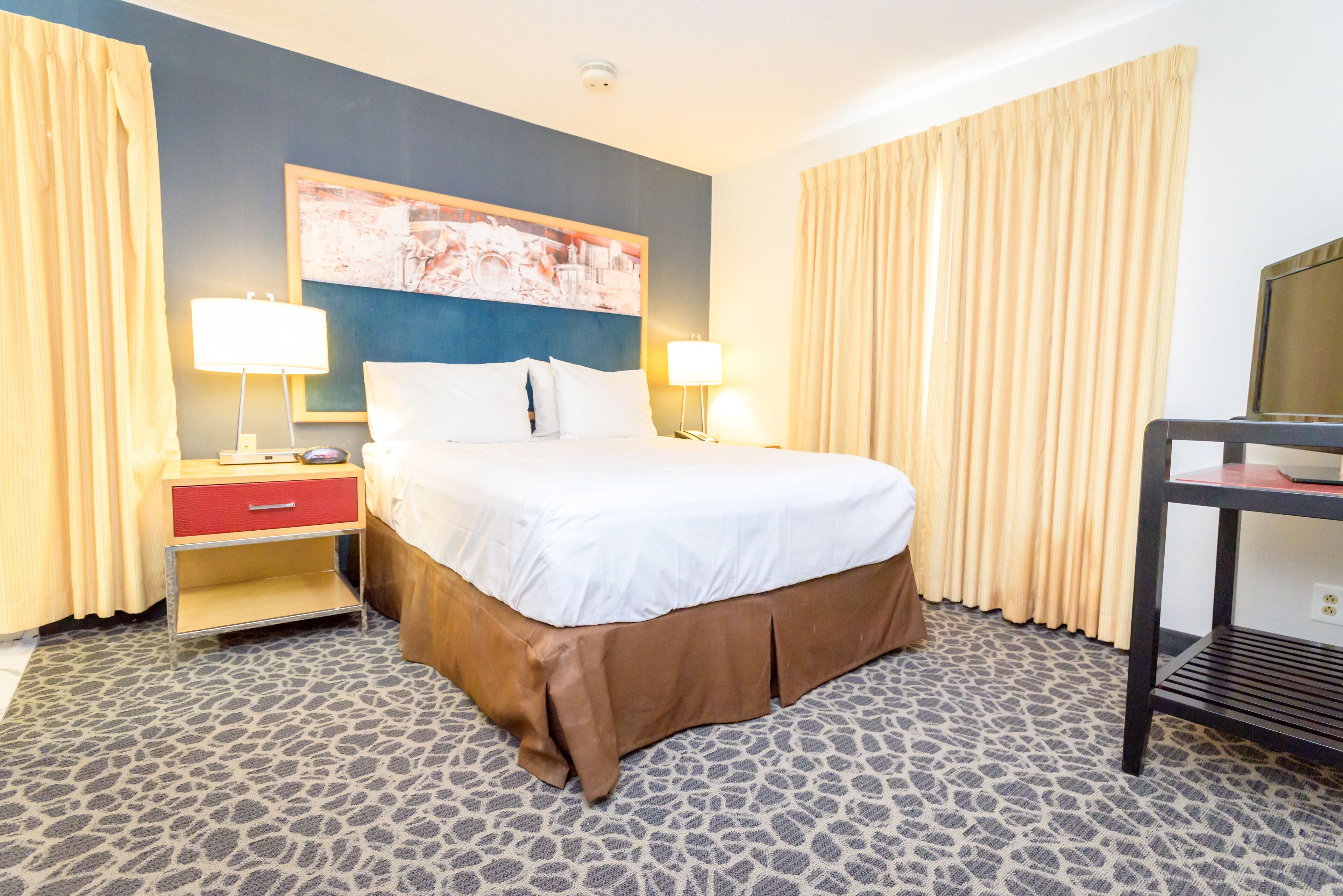 A hotel room with a bed, two bedside lamps, a nightstand, a TV on a stand, and a patterned carpet.