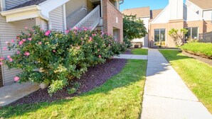 Un immeuble résidentiel doté d’un porche abrité, d’une façade en briques et d’un jardin bien entretenu agrémenté de fleurs roses.