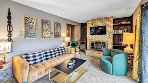 A living room with a sofa, armchair, coffee table, fireplace, bookshelf, and decorative wall art.