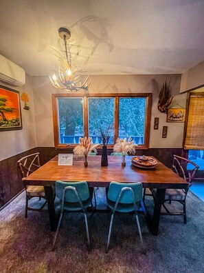 A dining area with a wooden table, four chairs, a chandelier, and a view of trees through large windows. - Woodland Cabin! Hot Tub, Arcade, Lake Access (Algoma)