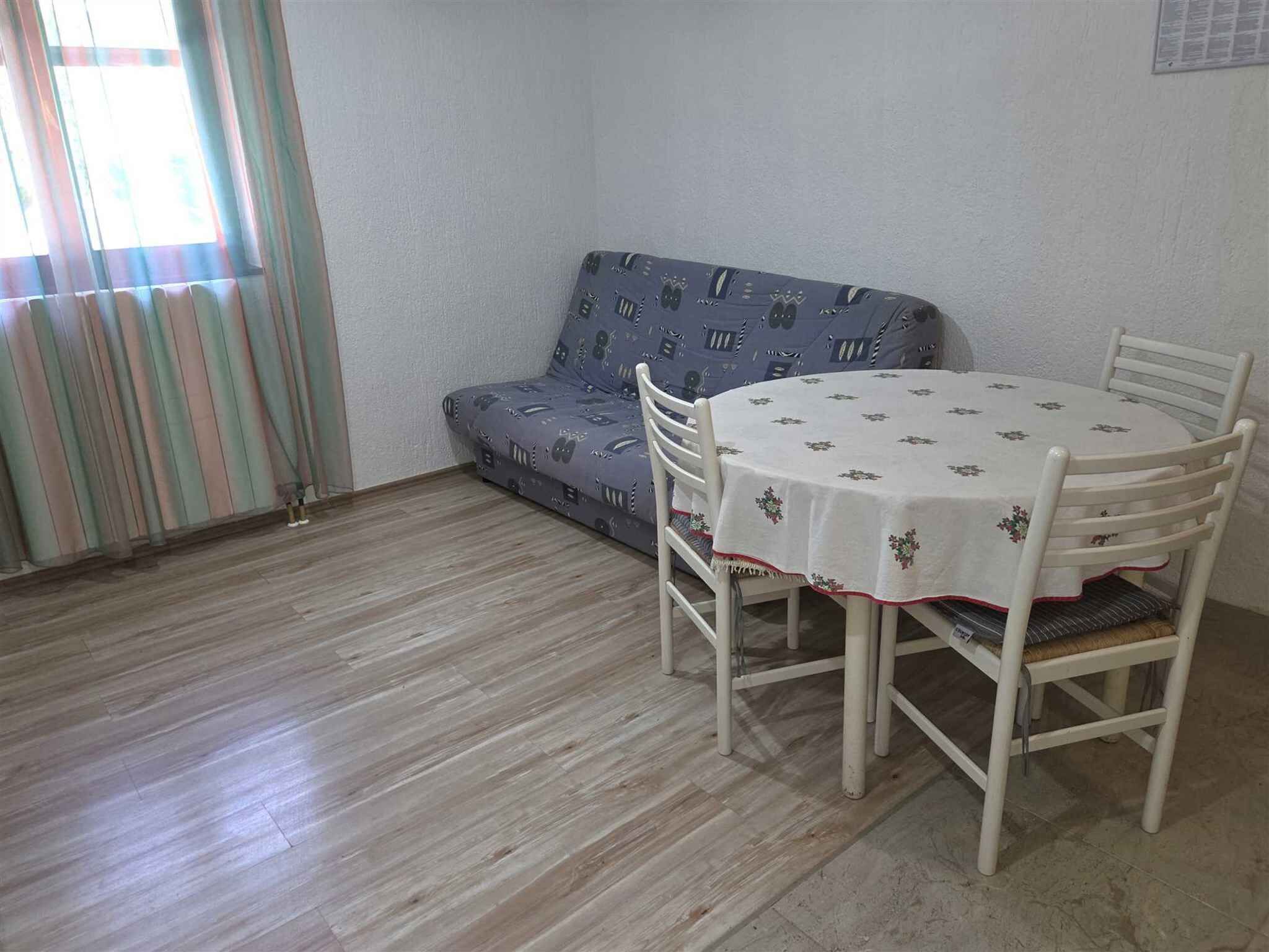Een kamer met een bank, een ronde tafel met stoelen, een raam met gordijnen en een lichtschakelaar aan de muur.