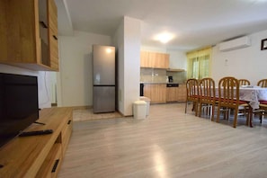 A compact living space with a kitchenette, a dining area with wooden chairs, a TV on a wooden stand, and a small white stool.