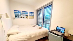 A bedroom with a bed, a desk with a laptop, a window with blinds, and a wall-mounted lamp. - Luxury 1 bedroom with Parking (Luxembourg City)