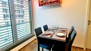 A dining area with a table set for four, a fireplace with a fire, and a large window with blinds. - Luxury 1 bedroom with Parking (Luxembourg City)