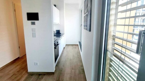 A narrow hallway with wooden flooring, white walls, and a large window with blinds. - Luxury 1 bedroom with Parking (Luxembourg City)