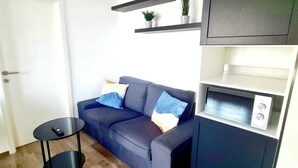 A modern living room with a dark grey sofa, two blue cushions, a small black side table, and a built-in microwave. - Luxury 1 bedroom with Parking (Luxembourg City)