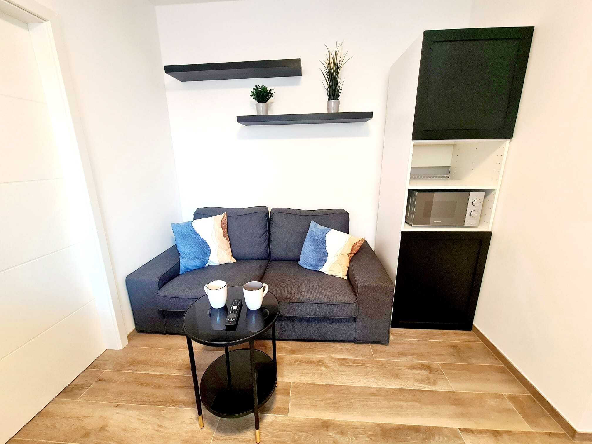 A modern living room with a grey sofa, two coffee cups, a remote control, and a small round table. There are two potted plants on floating shelves above the sofa.