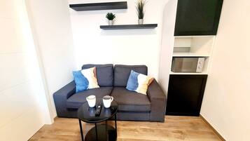 A modern living room with a grey sofa, two coffee cups, a remote control, and a small round table. There are two potted plants on floating shelves above the sofa.