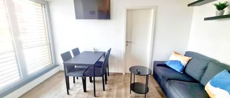 Uma sala de estar moderna com uma mesa de jantar, cadeiras, um sofá, uma TV fixada na parede e uma porta.