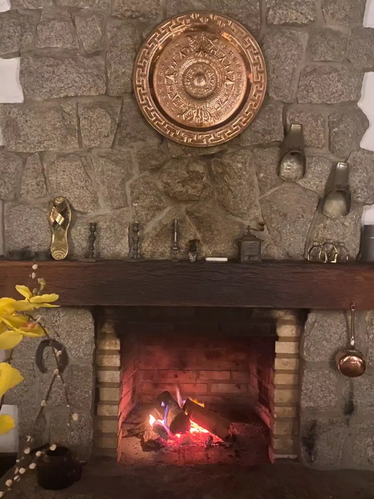 Ein Kamin aus Stein mit einer dekorativen Platte darüber, ein Feuer brennt darin, und verschiedene Gegenstände wie ein Kerzenhalter und ein Topf stehen auf der Kaminbank.