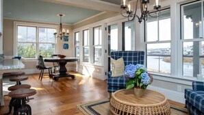 A spacious room with large windows, a dining area, and a seating area with a wicker table and blue sofa. - The Sea Eagle - Luxury 5 BR Waterfront Home in Boothbay Harbor with Dock & Sauna (Boothbay Harbor)