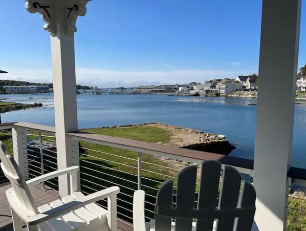 Une terrasse avec des chaises blanches donnant sur un lac paisible, d’où l’on aperçoit des maisons et un pont au loin.