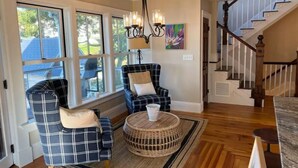 A spacious living room with a large window, a chandelier, a staircase, and a plaid sofa. - The Sea Eagle - Luxury 5 BR Waterfront Home in Boothbay Harbor with Dock & Sauna (Boothbay Harbor)