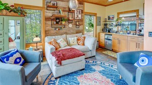 A cozy living room with a sofa, two armchairs, a coffee table, and a fireplace. There is a kitchen area with a refrigerator, stove, and sink. The room has wooden walls and a large window.