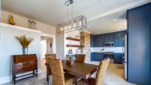 A modern dining room with a wooden table and wicker chairs, a kitchen with dark cabinets and stainless steel appliances, and a ceiling with exposed beams. - Sunset Bliss, Oceanview, Luxury, Hot Tub (Samoa)
