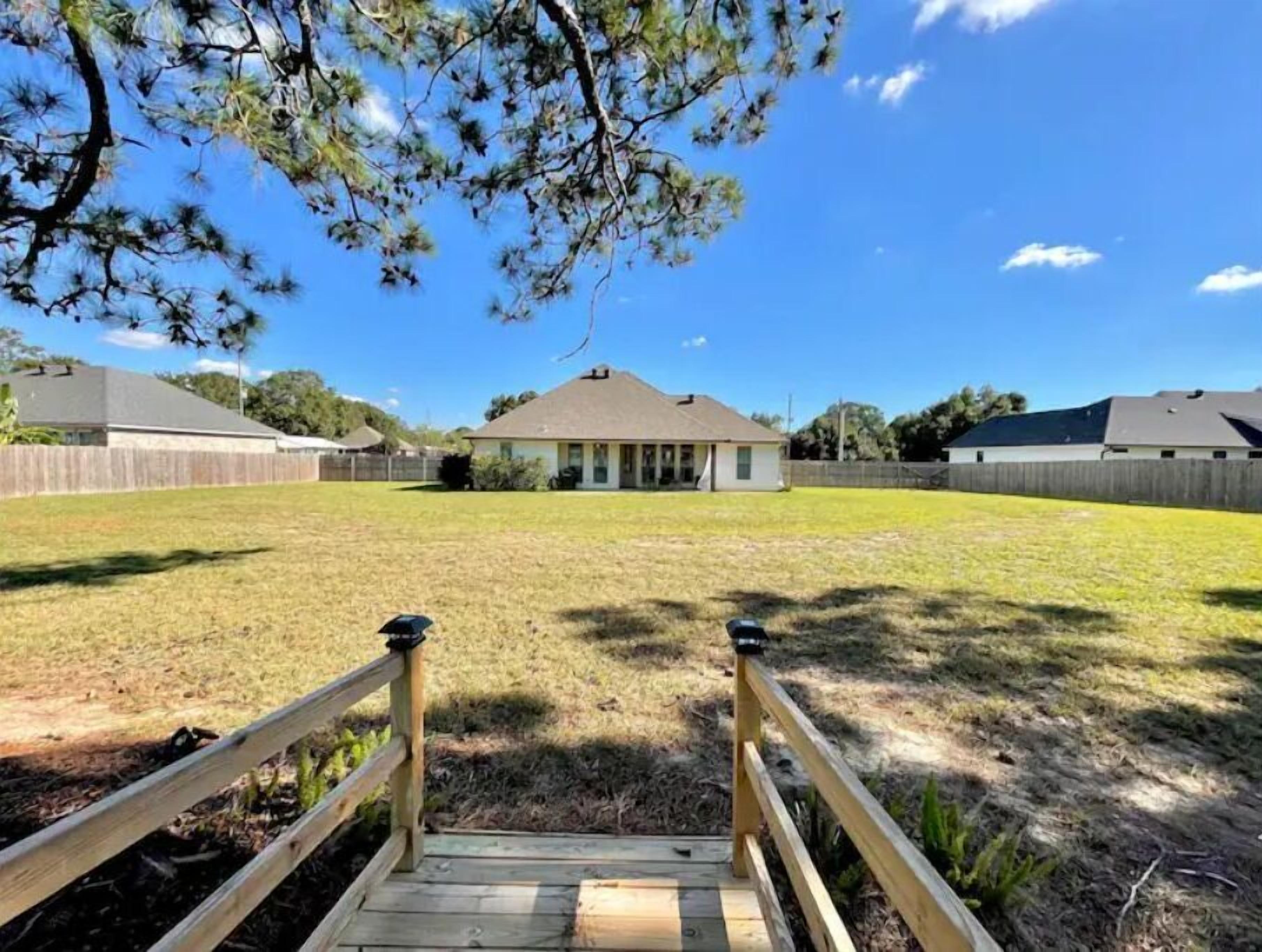 A backyard with a wooden deck leading to a house, surrounded by a fenced yard and a clear blue sky.