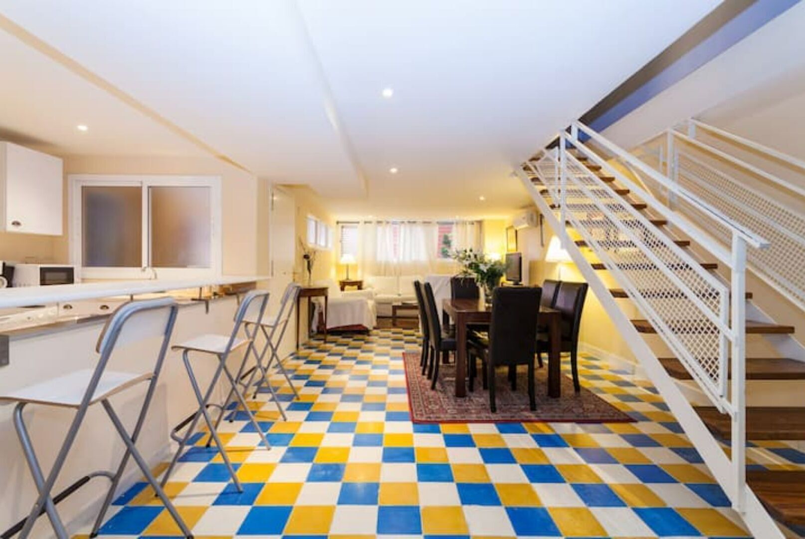 A hotel room with a checkered floor, a dining area with chairs and a table, a kitchen area with a counter and appliances, and a staircase with a railing.