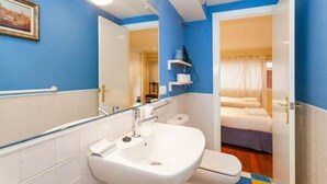 Une salle de bain avec un lavabo blanc, des toilettes et un espace douche. Un miroir est placé au-dessus du lavabo, et une porte donne accès à une chambre à coucher équipée d’un lit et d’une fenêtre.