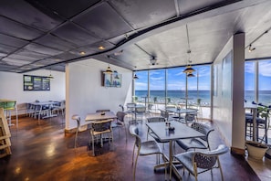 An interior with tables and chairs, a ceiling with lights, and a view of the ocean through large windows.