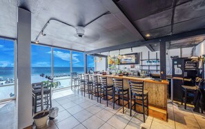 A bar area with a view of the beach, a bar counter, and several chairs.