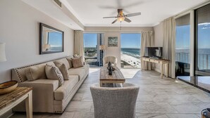 A spacious living room with a large window offering a view of the beach, a ceiling fan, and a flat-screen TV on a stand.