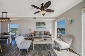 A modern living room with a ceiling fan, a dining area with a view, and a large window. - Tidewater1418 Corner Views WOW, FREE Golf&Perks, Pools, Hot Tubs by Pier Park (Panama City Beach)