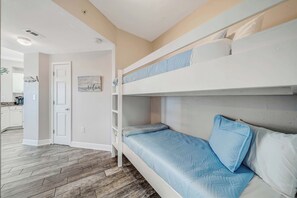 Una cama tipo litera con colchón y almohadas azules, una puerta blanca y una cocina al fondo.
