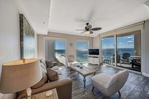 A spacious living area with a large window overlooking a beach, a ceiling fan, and a television on a stand. - Tidewater1418 Corner Views WOW, FREE Golf&Perks, Pools, Hot Tubs by Pier Park (Panama City Beach)