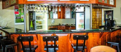 Zona de bar con un letrero de Castle Lager, un letrero de Miller Genuine Draft y un letrero de Peroni. Hay tres taburetes y una barra con un refrigerador y estantes llenos de botellas.