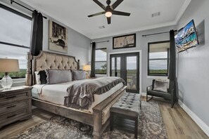 A bedroom with a large bed, a ceiling fan, a window, and a TV on the wall.