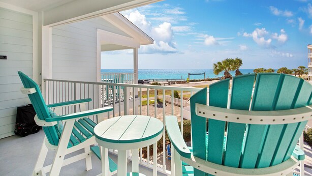 Un balcon agrémenté de chaises Adirondack turquoise et d’une petite table ronde, donnant sur une plage bordée de palmiers et surplombant un ciel bleu azur.