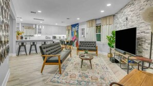 Una sala de estar moderna con un sofá, sillones, una mesa de centro y un televisor. Se observa una cocina al fondo con taburetes de bar y una zona de comedor.