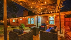 Un patio al aire libre con mobiliario de mimbre, luces colgantes y un edificio de ladrillo al fondo.