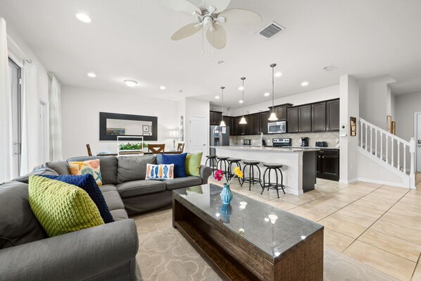 A modern living room with a gray sofa, a glass coffee table, and a kitchen area with dark cabinets and a white island. - Gorgeous 4Bd TH Pool Windsor at Westside-2018WW (Kissimmee)