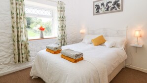 A bedroom with a bed, two bedside lamps, a window with curtains, a painting on the wall, and a vase with flowers on the windowsill. - Coed y Glyn Bach Equestrian (Caernarfon)