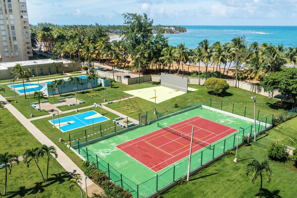 Vista aérea de un complejo deportivo con varias canchas de tenis, una piscina y una playa al fondo.